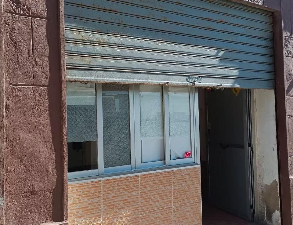 Persiana metálica cerrada sobre el escaparate de una tienda con una pequeña ventana y una puerta en una fachada de ladrillo.