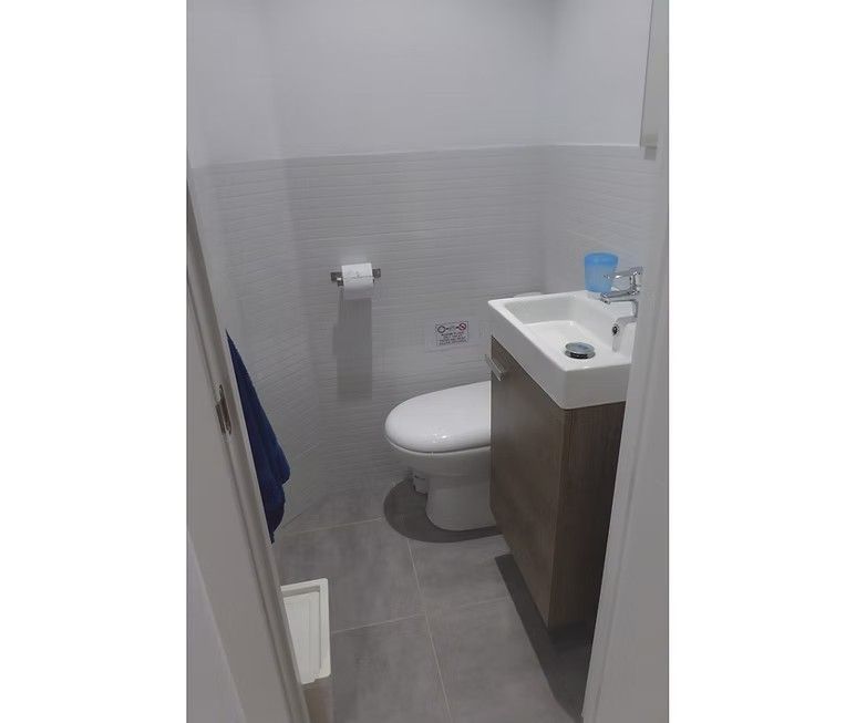 Baño pequeño con inodoro, lavabo con mueble, paredes de azulejos grises y una taza azul sobre la encimera.