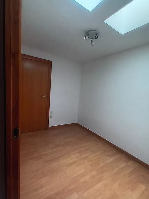 Habitación blanca vacía con suelo de madera, puerta y dos ventanas tipo tragaluz.