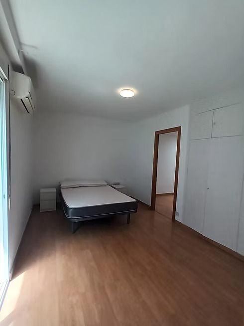 Habitación vacía con una cama, suelo de madera, paredes blancas y una puerta abierta.