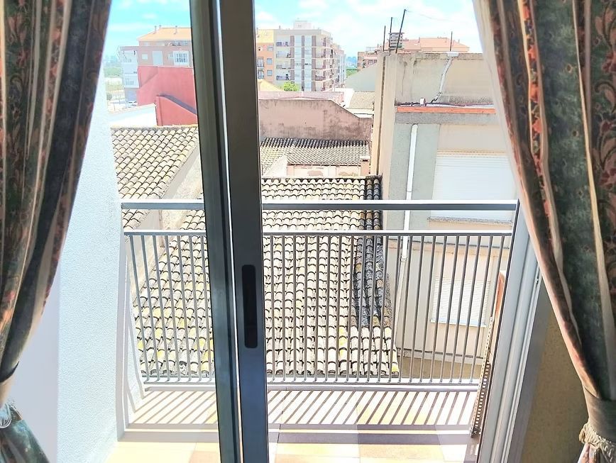 Puerta corrediza de cristal que da a un balcón con barandilla metálica, cortinas y vistas a los tejados del exterior.