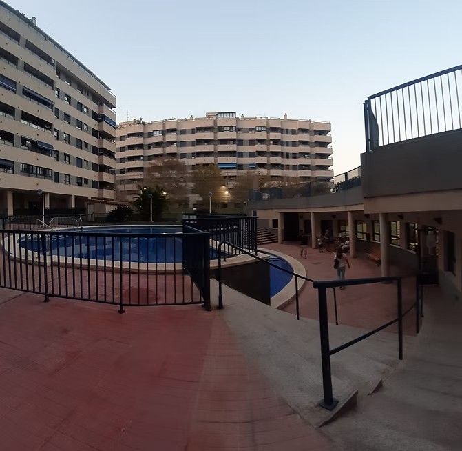 Patio de apartamentos con piscina azul, pasarela curva y edificios de varias plantas al atardecer.
