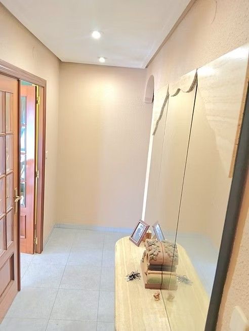Pasillo estrecho de color beige con suelo de baldosas, puerta de madera abierta y un pequeño banco con objetos decorativos.