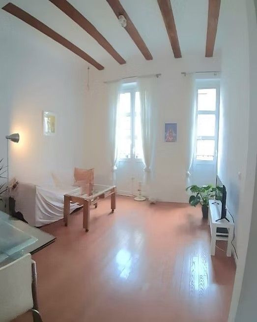 Sala de estar minimalista y luminosa con paredes blancas, vigas de madera, sofá, silla, ventanas y una planta en maceta.