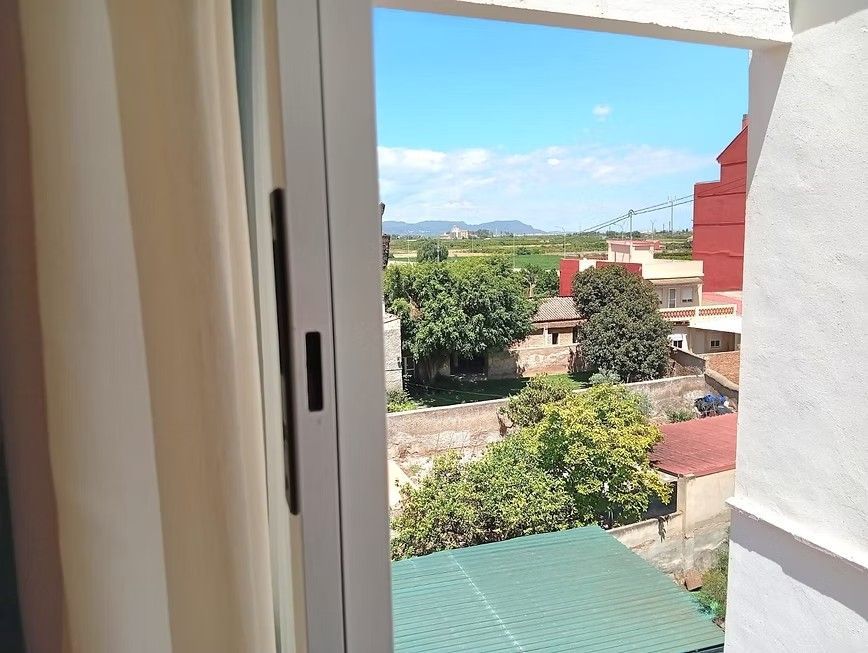 Vista a través de una ventana abierta de árboles, tejados y un cielo azul en un día soleado.