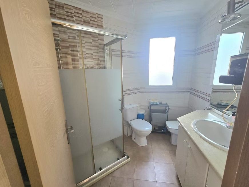 Baño pequeño con inodoro, ducha, lavabo, azulejos beige y ventana esmerilada.