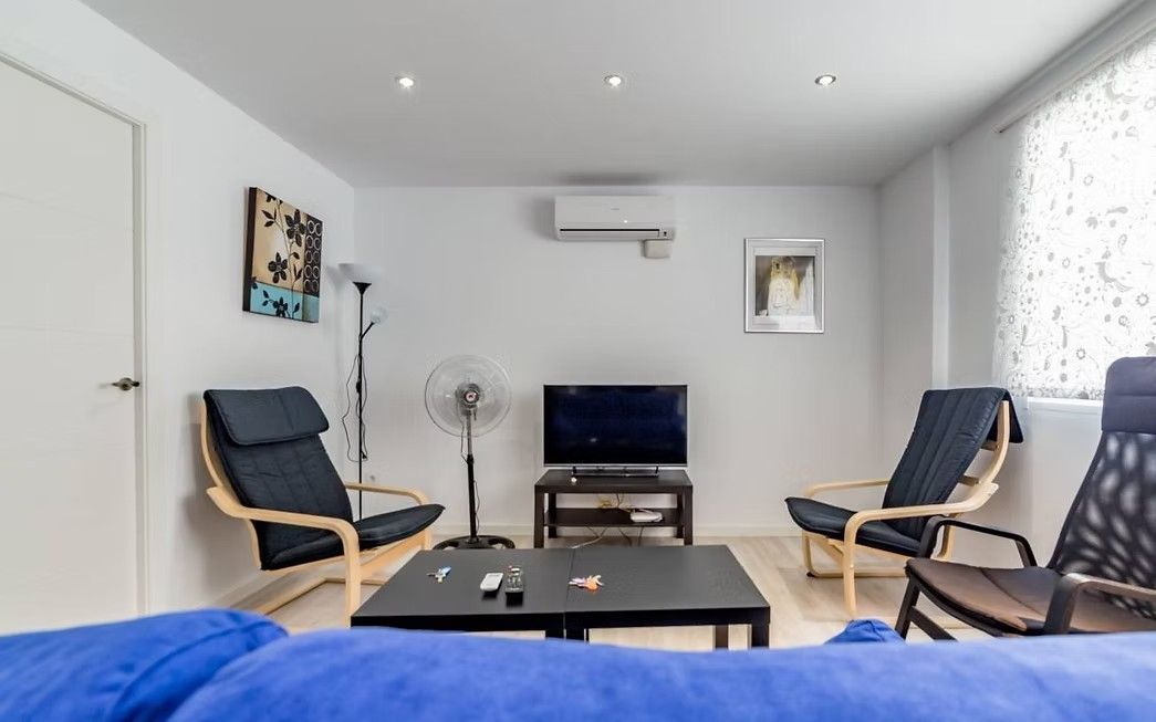 Sala de estar moderna con sillas negras, una mesa de centro baja, televisor, lámpara de pie y borde de sofá azul.