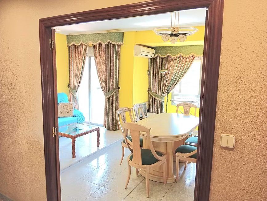 Comedor con paredes amarillas, mesa redonda, sillas y cortinas estampadas, visto a través de una puerta.