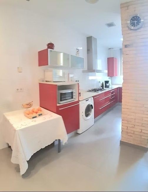 Cocina luminosa y compacta con armarios rojos, electrodomésticos blancos, una mesa y un reloj de pared.