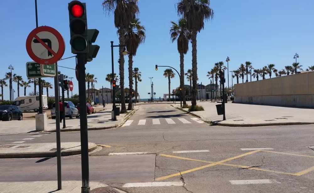 Intersección soleada con semáforo en rojo, palmeras, paso de peatones y señal de prohibido girar a la izquierda.