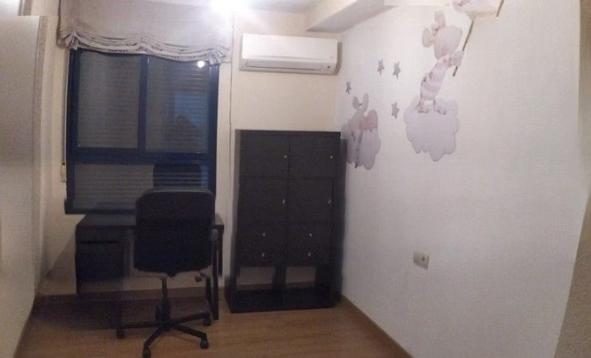 Habitación pequeña con un escritorio, una silla negra, aire acondicionado y un armario oscuro junto a la ventana.
