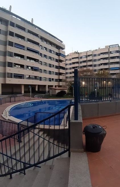 Zona de piscina del complejo de apartamentos con agua azul, barandillas negras y altos edificios beige al atardecer.
