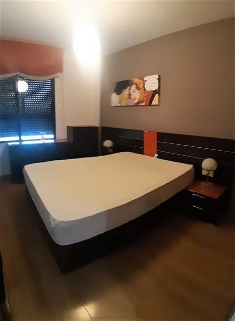 Dormitorio moderno con una cama grande, suelo de madera oscura, persianas y cuadros en la pared.
