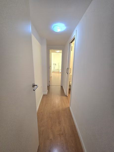 Un estrecho pasillo blanco con suelo de madera y una lámpara de techo azul, que conduce a una puerta luminosa.