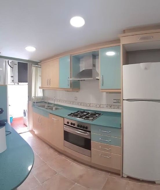 Cocina moderna con gabinetes de madera clara, encimeras turquesa, refrigerador blanco y estufa de gas.