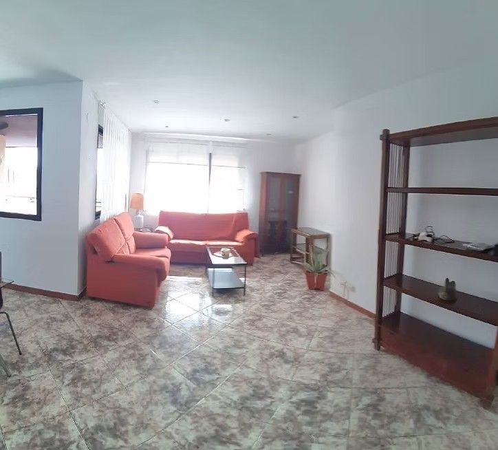 Luminoso salón con sofás rojos, suelo de baldosas y una estantería de madera junto a una gran ventana.