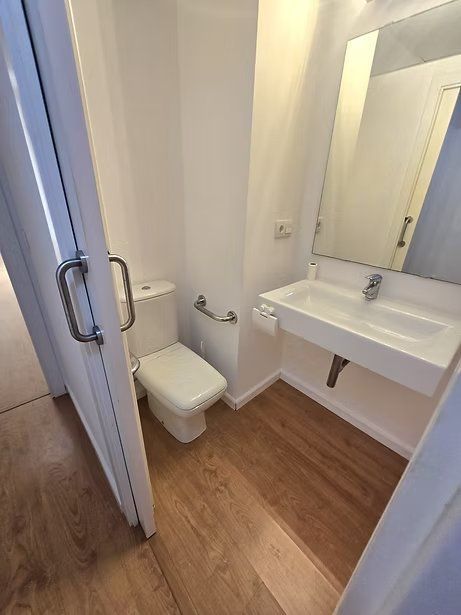 Baño pequeño y accesible con inodoro, lavabo mural, espejo y suelo con aspecto de madera.