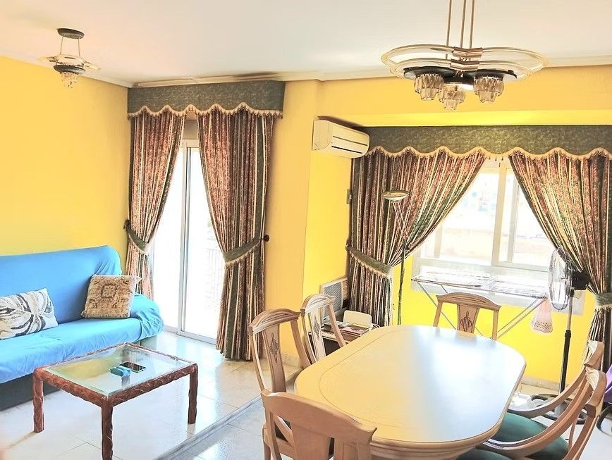 Luminoso salón-comedor con sofá azul, mesa de comedor, paredes amarillas y grandes ventanas con cortinas.