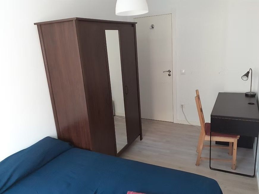 Habitación pequeña con cama azul, armario de madera, escritorio, silla y puerta blanca.