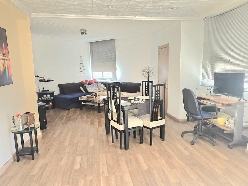 Pequeña sala de estar con un sofá negro, mesa de comedor, escritorio y suelo de madera, bañada por la luz natural.