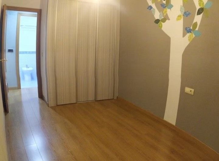 Habitación vacía con suelo de madera, paredes beige, puertas de armario y una puerta abierta que da a un baño luminoso.
