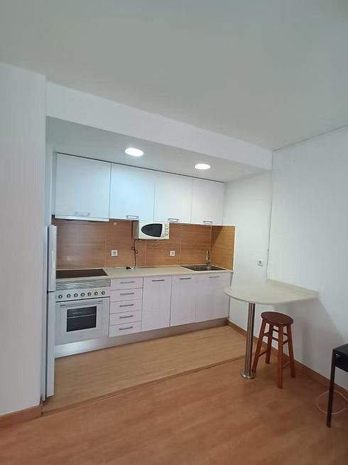 Pequeña cocina blanca con suelo de madera, fogones, fregadero, armarios y una mesita con taburete.