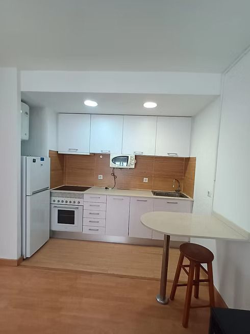 Pequeña cocina blanca con suelo de madera, armarios, fogones, nevera y una mesa redonda con taburete.