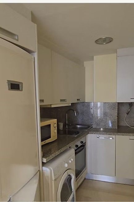 Cocina compacta blanca con nevera, lavadora, microondas y armarios de esquina con salpicadero oscuro.