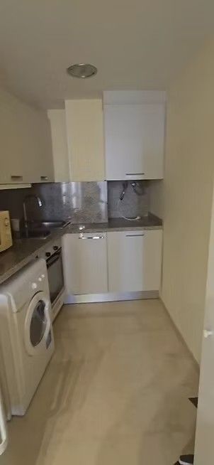 Cocina estrecha tipo galera con gabinetes blancos, salpicadero gris, lavadora y suelo de baldosas beige.