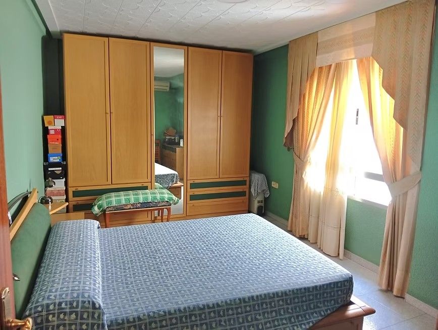Habitación pequeña con cama, armarios de madera y luz natural que entra por una ventana con cortinas.