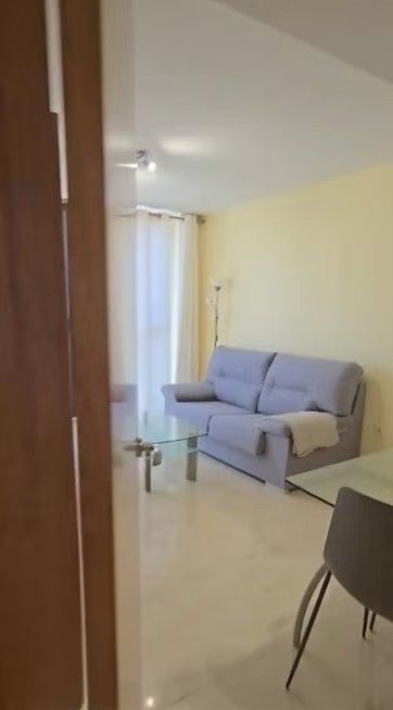 Sala de estar luminosa con un sofá gris, una mesa de centro de cristal y una silla de comedor cerca de una puerta.