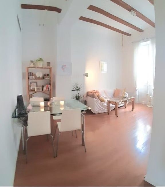Luminoso salón con paredes blancas, vigas de madera, sofá, mesa de comedor y luz natural que entra por un gran ventanal.