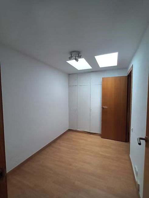 Habitación blanca vacía con suelo de madera, armario empotrado y puerta de madera abierta bajo luces de techo.