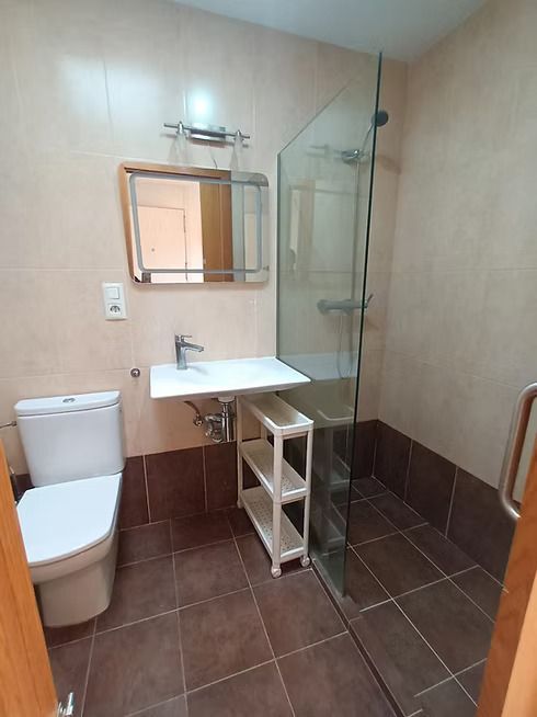 Pequeño baño de color beige con inodoro, lavabo, espejo y cabina de ducha de cristal sobre suelo de baldosas marrones.