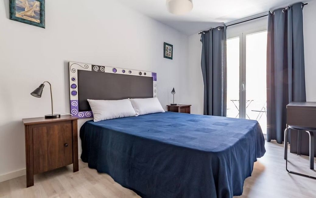 Habitación luminosa con colcha azul, mesita de noche de madera y puertas correderas de cristal con cortinas.