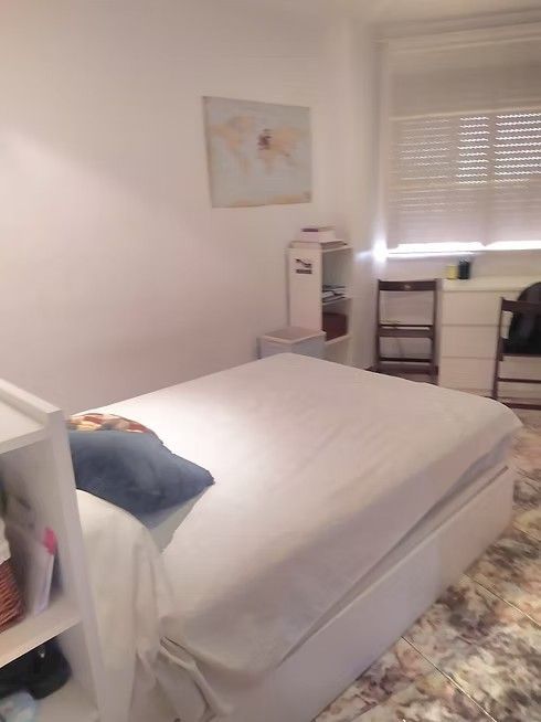 Habitación pequeña con cama, almohada azul, escritorio, silla, persianas y cuadros en la pared.