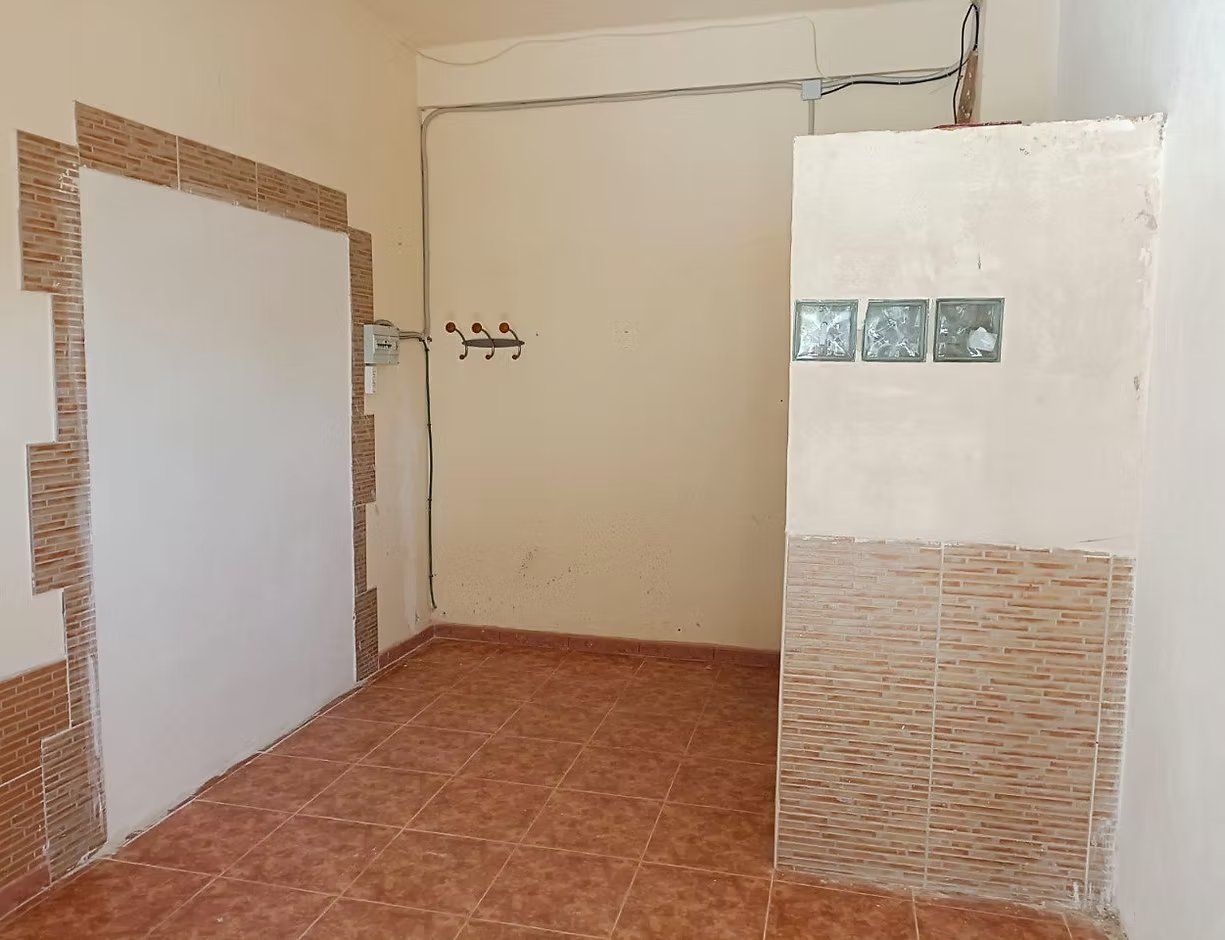 Habitación vacía con azulejos, paredes de color beige, una puerta blanca y pequeños bloques de vidrio en la pared derecha.
