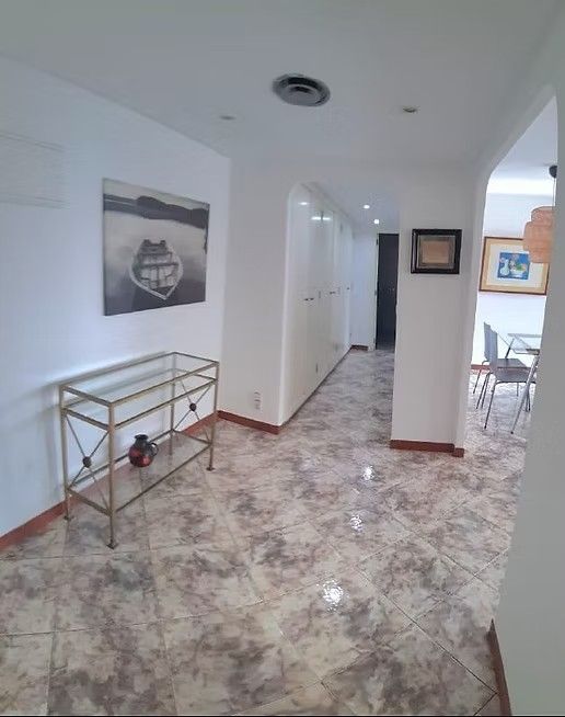 Pasillo luminoso con paredes blancas, suelo de baldosas estampadas y una pequeña mesa de metal contra la pared izquierda.
