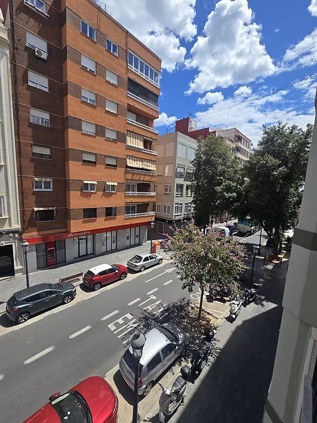 Calle soleada de la ciudad con coches aparcados, un edificio de apartamentos naranja y árboles frondosos bajo un cielo azul.