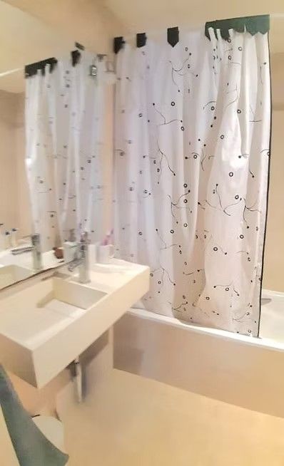 Baño pequeño con lavabo y bañera blancos, cortina de ducha blanca con puntos negros y suelo de baldosas beige.