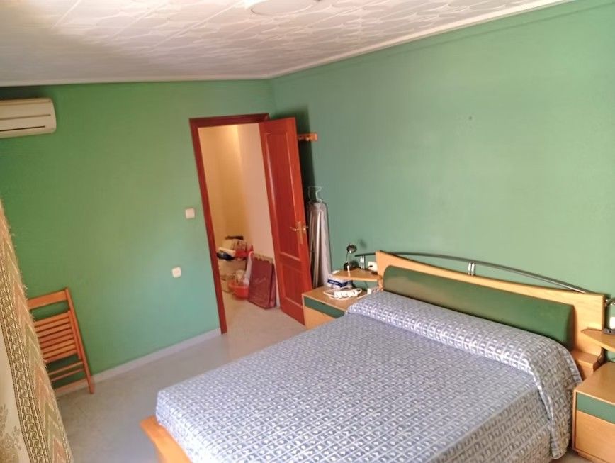 Habitación sencilla con una cama, paredes verdes y una puerta abierta que da a un pasillo.