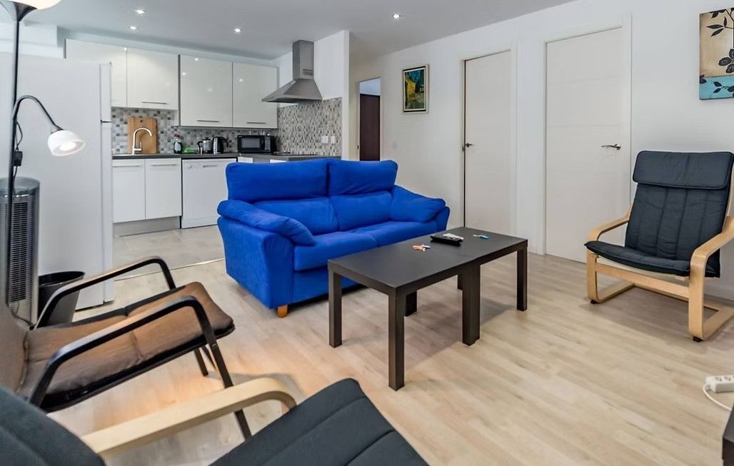Sala de estar moderna con un sofá azul brillante, una mesa de centro negra y sillas de madera en una zona de cocina abierta.