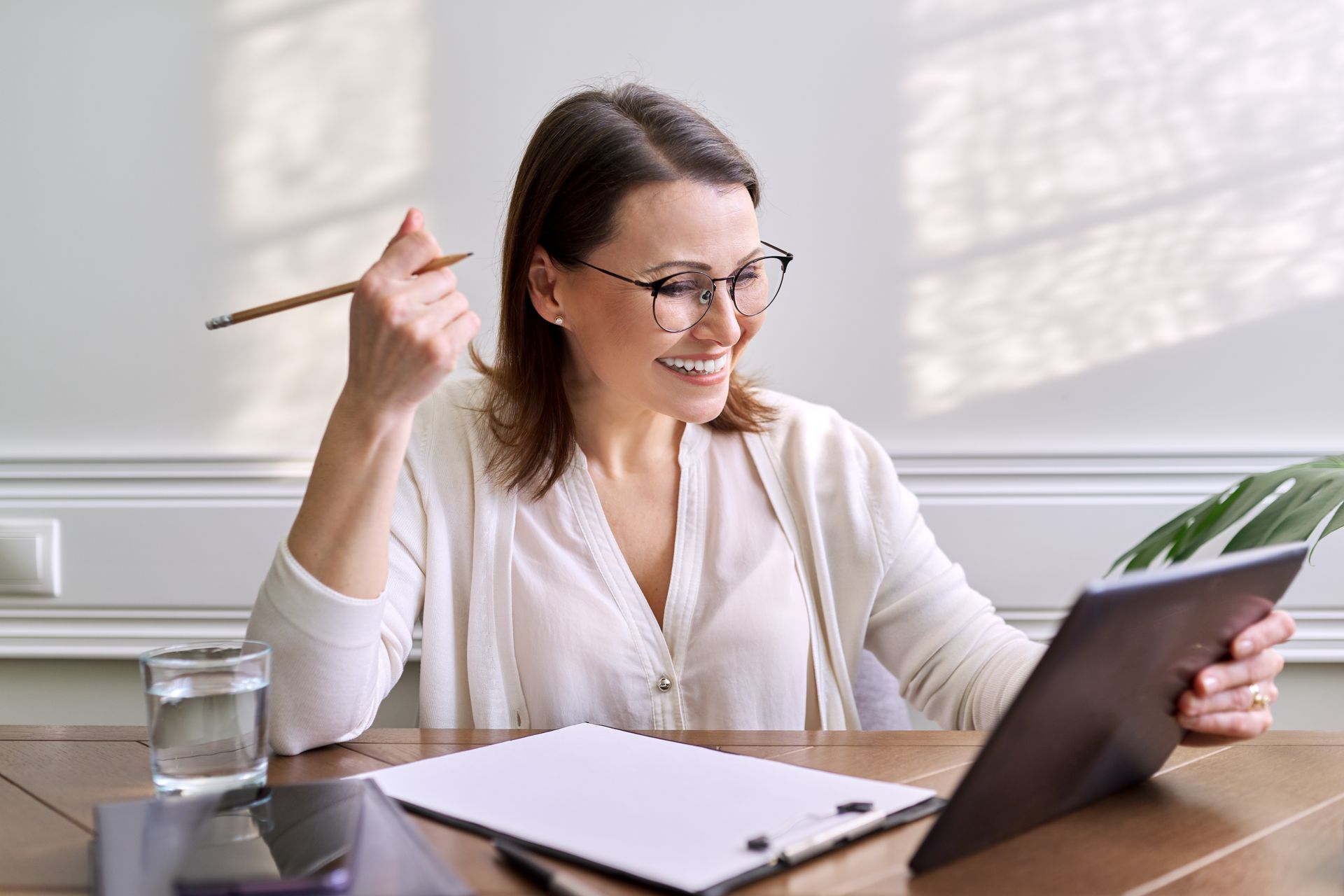 Une femme à lunettes sourit en regardant une tablette, un crayon à la main. À côté d'elle se trouvent un verre d'eau et un bloc-notes.