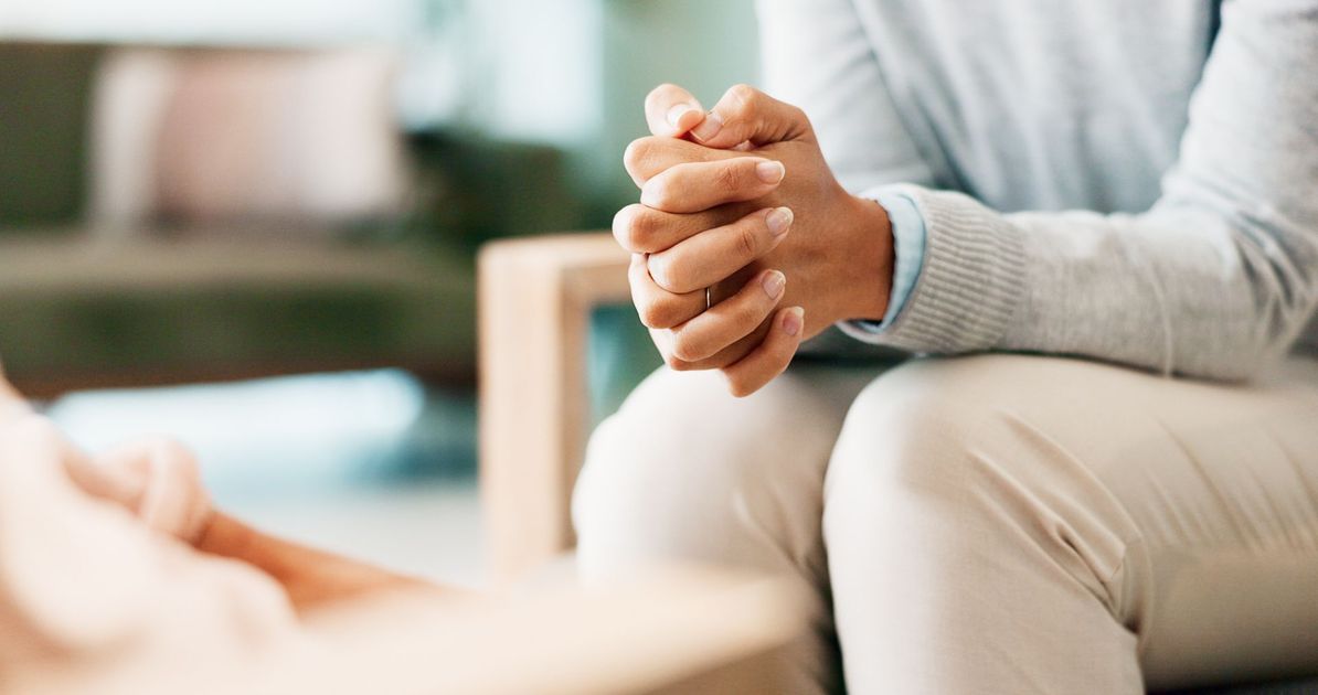 Personne assise, les mains jointes, en séance de thérapie ; éclairage tamisé, en intérieur.