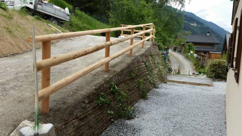 Chemin de gravier rural longeant un muret en pierre incliné avec une rambarde en bois.