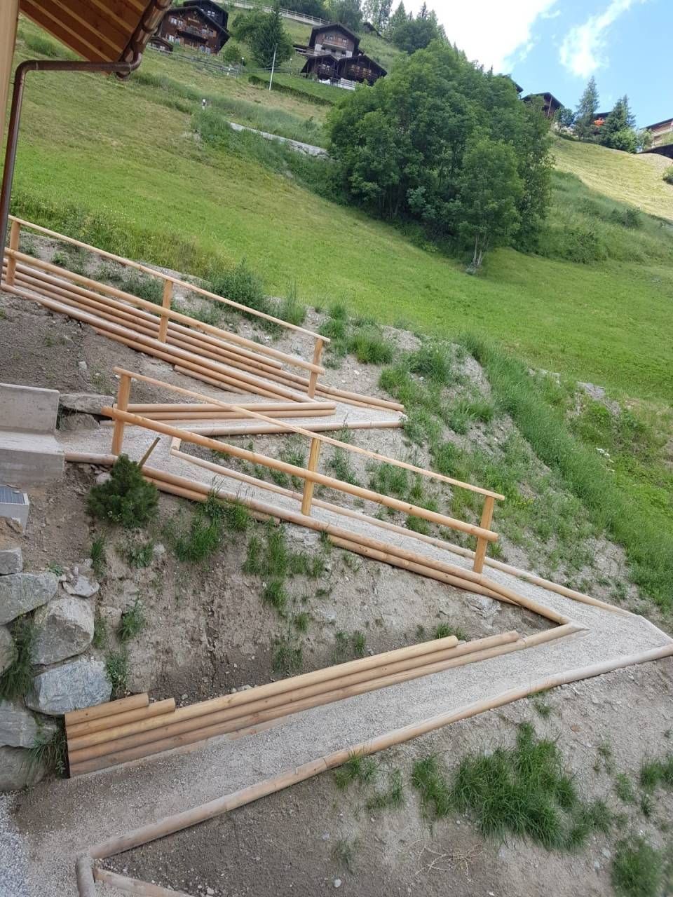 Pont en bois avec rambardes sur un sentier de montagne, à flanc de colline herbeuse et rocheuse.
