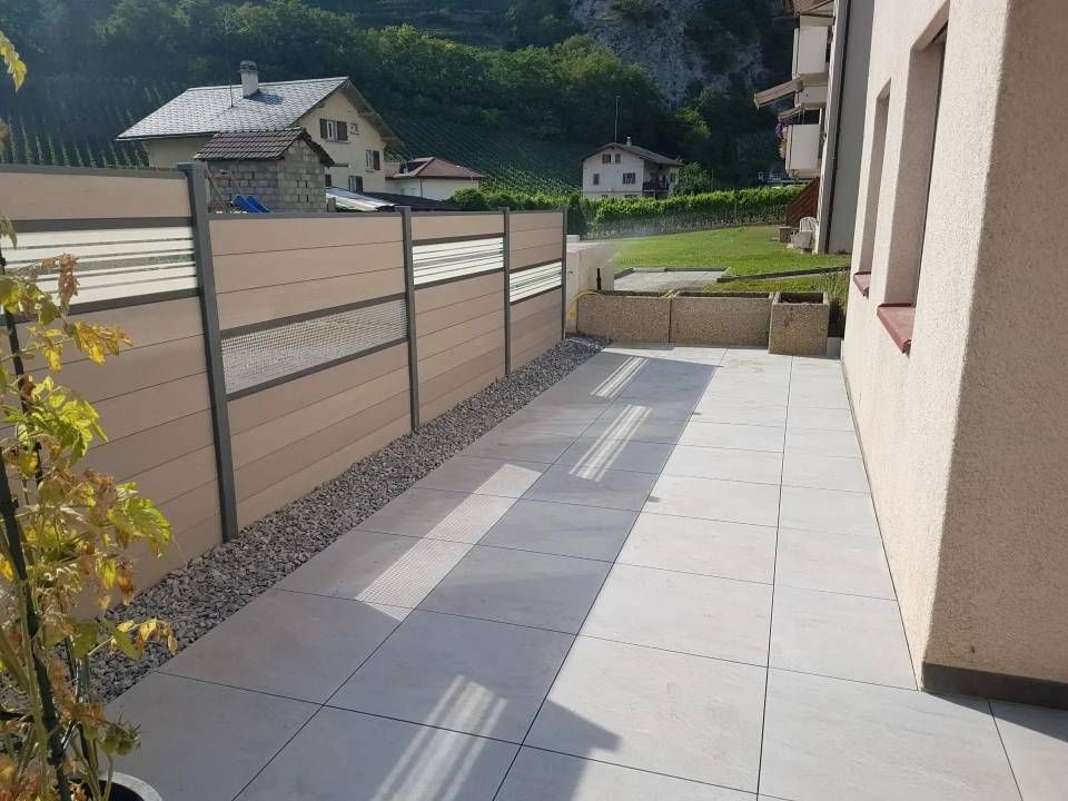 Cour latérale étroite et pavée jouxtant une maison, avec une clôture, une bordure de gravier et une zone herbeuse devant.