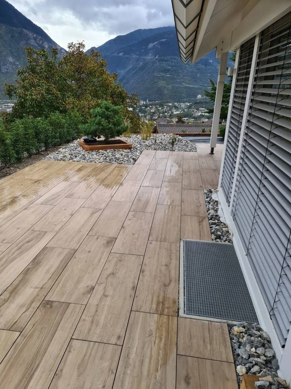 Terrasse en bois donnant sur les montagnes et un village au bord d'un lac, à côté d'une maison aux volets gris.