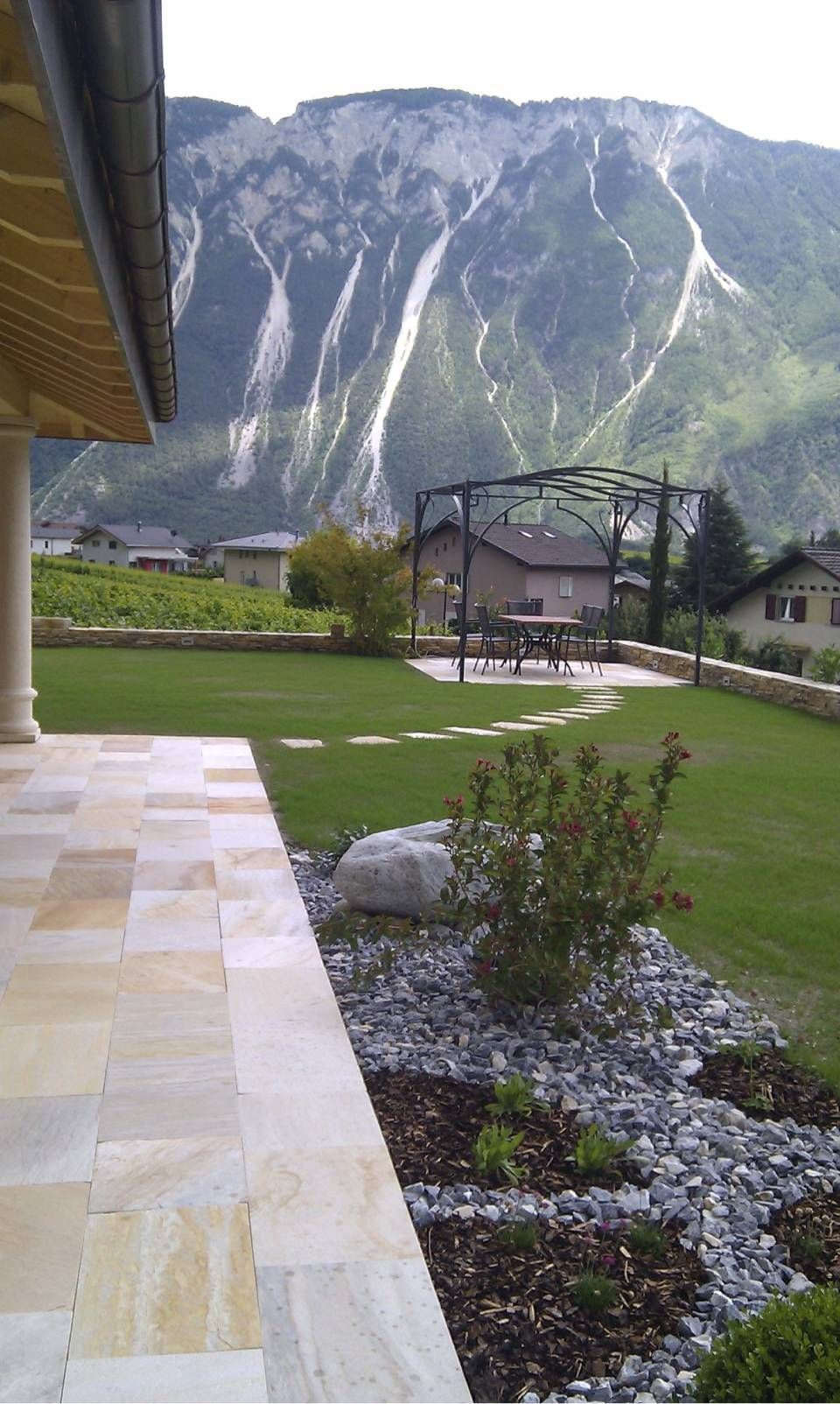 Cour intérieure d'un chalet de montagne avec pelouse verte, chemin de pierre et falaises.