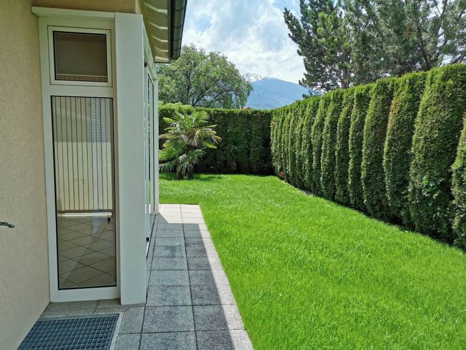 Terrasse arrière attenante à une maison, avec pelouse verte, hautes haies et montagnes à l'horizon.
